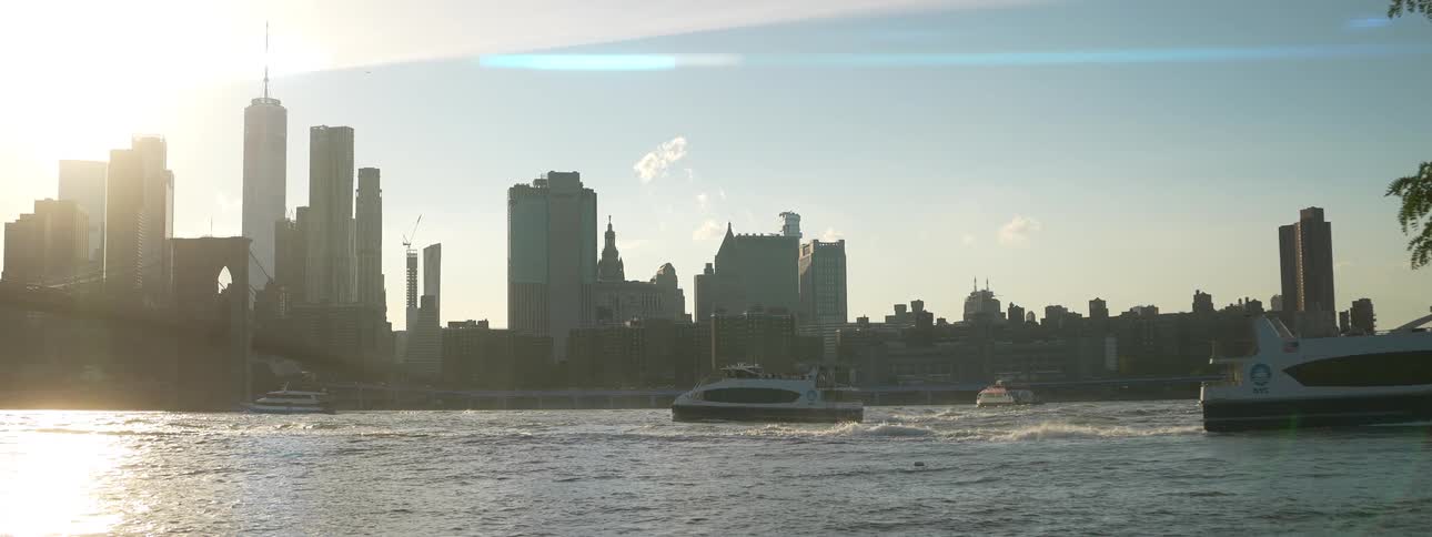 A modern white and blue ferry boat is traveling across a wide choppy waterway leaving a visible wake behind it as it moves past the silhouetted skyline of a major city which includes several towering skyscrapers and other buildings