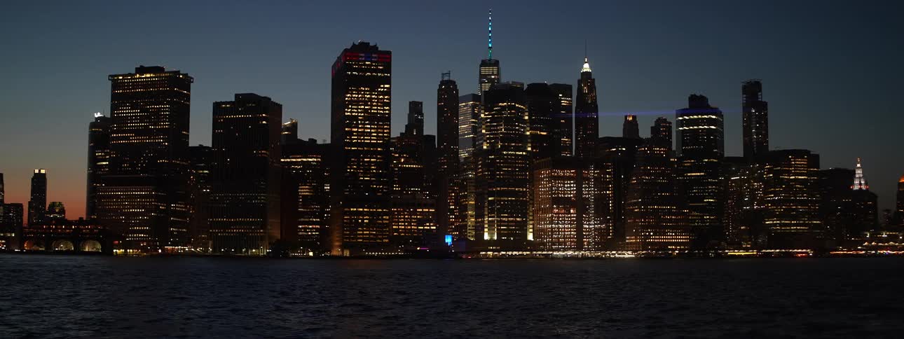 A major metropolitan area is showcased at night with numerous tall skyscrapers dominating the frame many of their windows brightly lit with goldenyellow light contrasting sharply against the deep blue twilight sky and the dark water in the foreground