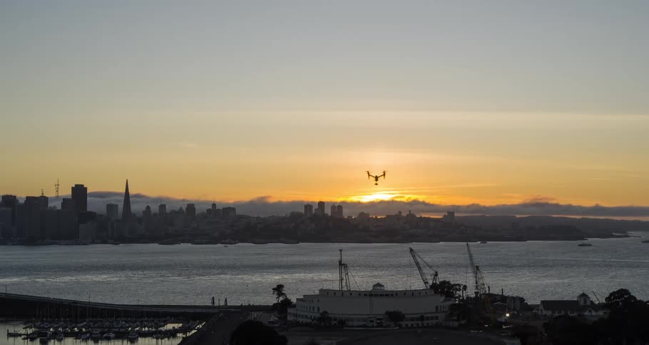 A small drone is hovering in the sky silhouetted against a bright sunset with the dark outline of a major city skyline stretching across the horizon behind a body of water and a marina and construction site visible in the dark foreground
