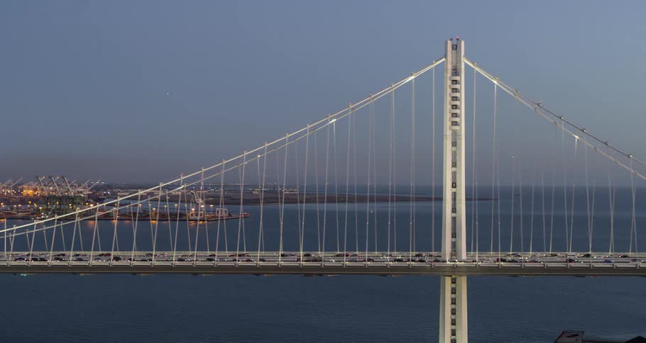 A modern suspension bridge is centered over a wide expanse of dark bay water with the stream of traffic lights visible on its roadway while the illuminated tower and cables stand out against the blue sky with the lights of a distant city visible