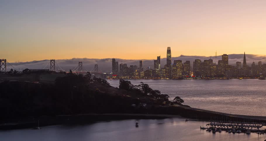 A large city with skyscrapers and a large bridge spanning a vast body of water on an orangesky morning