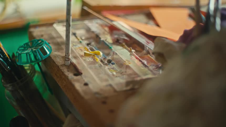 A closeup shot shows a segment of a wooden palette covered in various streaks of brightly colored paint with paint brushes in a container nearby