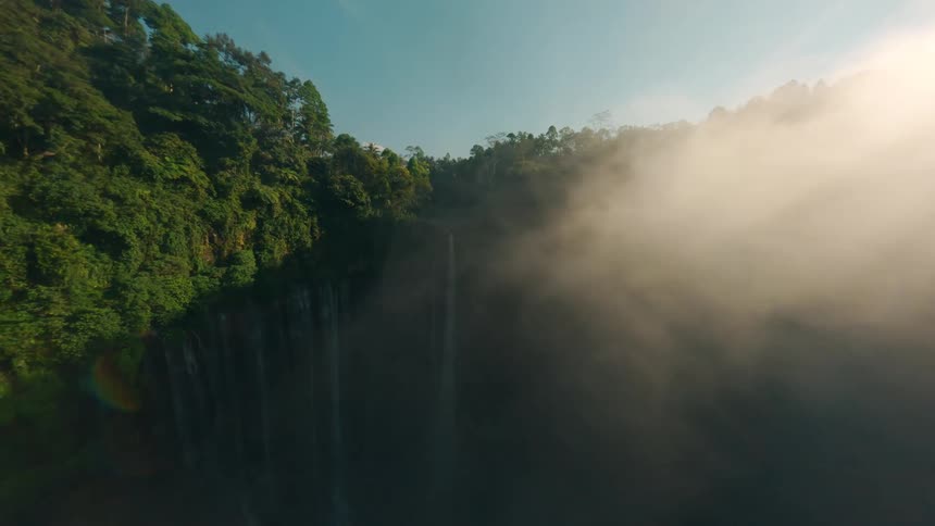 Light glare and mist obscure a rainforest and the waterfalls flowing down a cliff on a sunny day