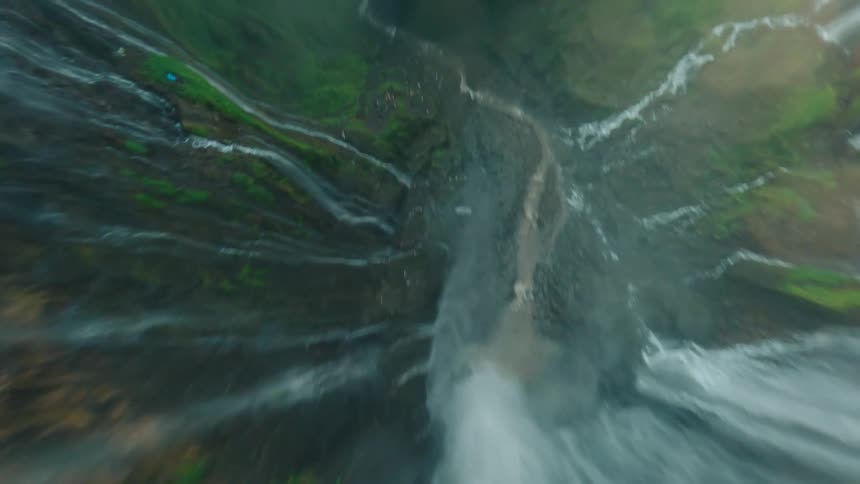 A drone footage that descend rapidly over the edge of a tall multitiered waterfall revealing the sheer mossy rock faces of the surrounding gorge and the lush green jungle canopy above