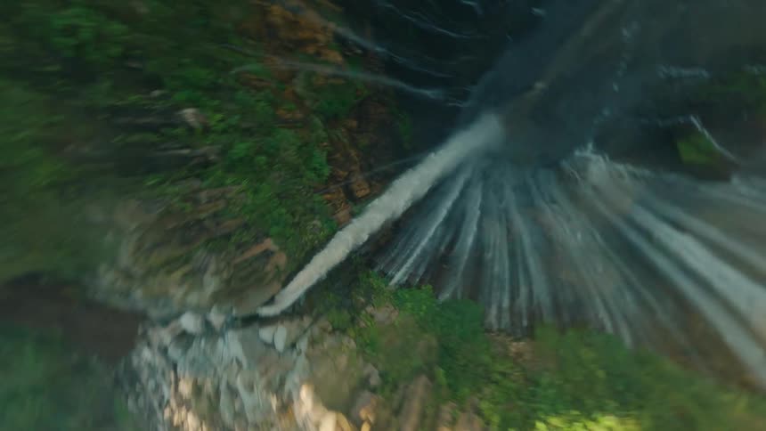 A drone view of a waterfall showcasing the tremendous height and the vertical drop into a deep shadowed gorge with the surrounding cliffs covered in dense green jungle and bright sunlight while people hiking on the rocks