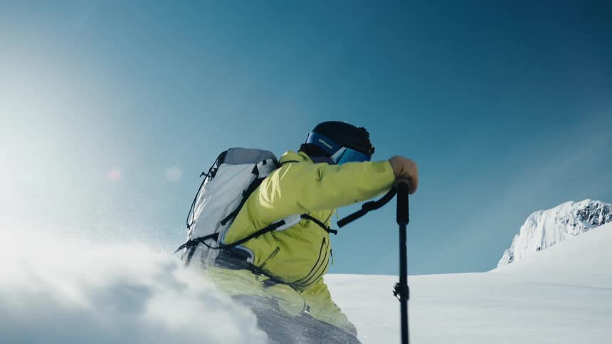 A skier wearing a bright yellow jacket speeds down a massive snowcovered mountain slope carving a turn that sprays a thick cloud of white powder into the bright afternoon air