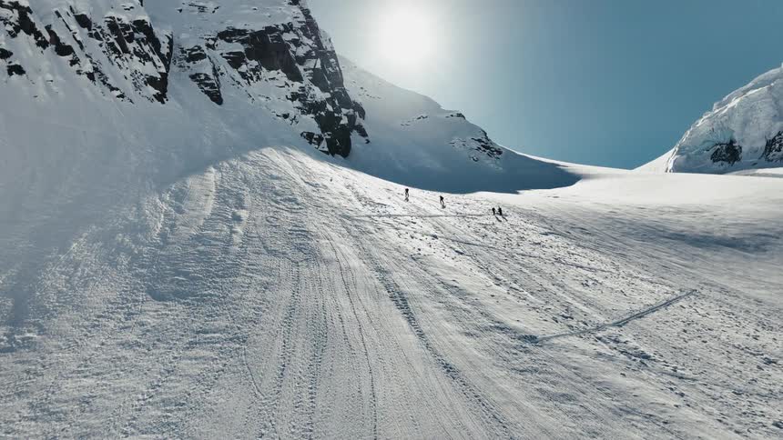 A group of snowboarders is descending a wide very steep snowfield covered in fresh tracks situated high in a sunny alpine environment between dark rocky cliffs and a bright glacier