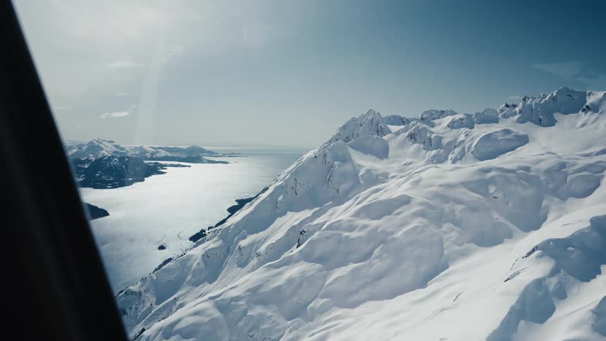 An aerial view from an aircraft window shows vast snowcovered mountains rising above a wide river or inlet on a clear day