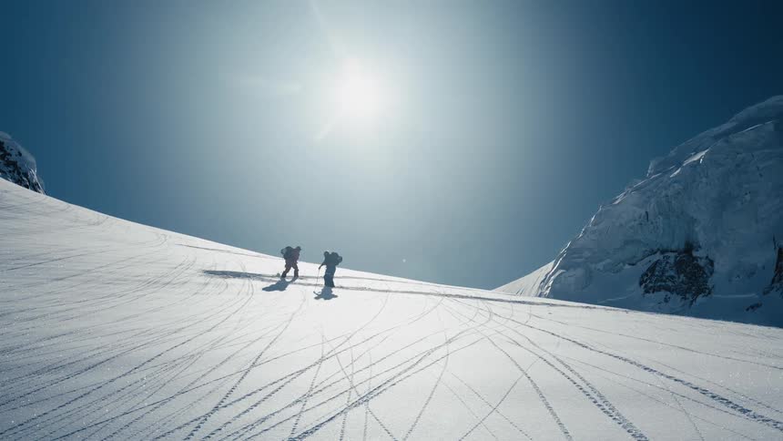 Two people on skis traverse across a vast steep snowcovered mountain slope against the bright sun with one subject passing the other as they slowly make their way across the white expanse