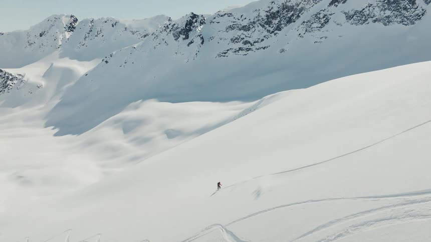 A lone snowboarder carves sweeping turns down a massive untouched snowcovered mountain their tiny figure trailing a long track across the vast alpine landscape