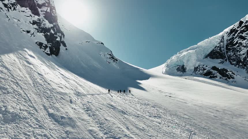 A group of skiers makes their way up a vast sun drenched mountain pass
