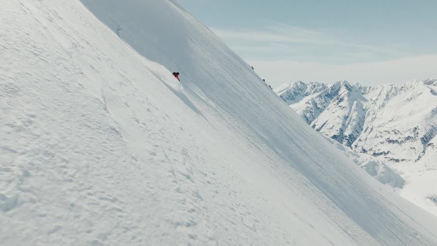 A lone snowboarder carves down a steep wide open mountain face with a vast snowy mountain range in the distance