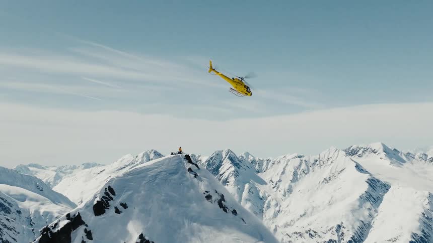 Yellow helicopter hovers above a snowy peak surrounded by mountains