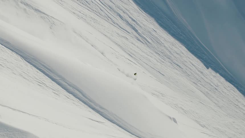 A snowboarder wearing a bright yellow jacket is aggressively descending a very steep vast snowfield executing turns that spray large plumes of powder into the air with the massive scale of the mountain face dominating the frame