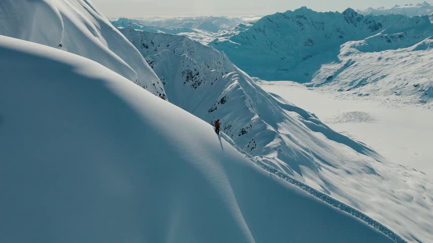 A lone skier stands on a steep snowcovered ridge surrounded by dramatic sunlit mountain peaks
