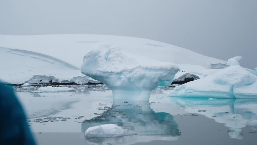 A blue iceberg drifts in dark water amid icy slopes under a pale sky