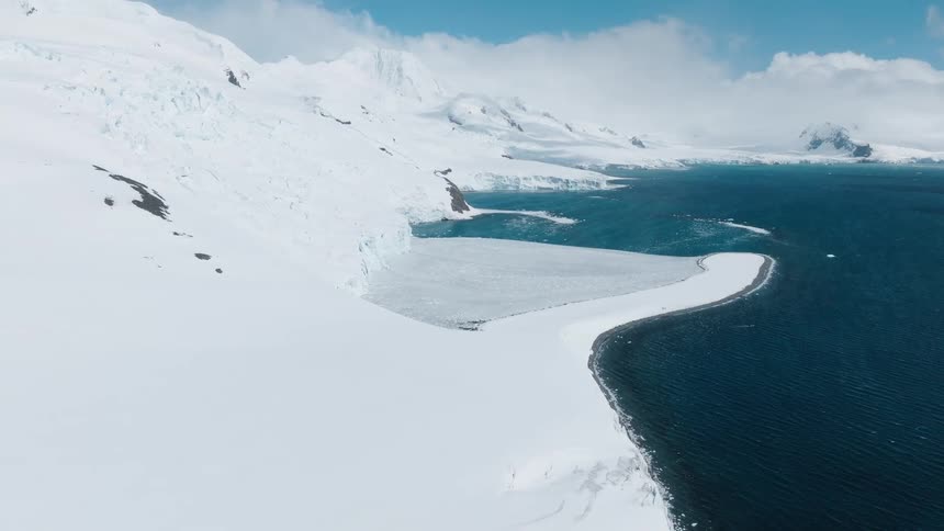 A sweeping view of a frozen coastline shows snowcovered mountains meeting the deep blue arctic sea under a bright sky