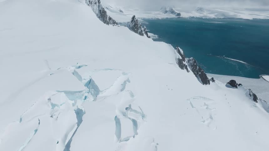 Vast snowy mountains stretch toward a turquoise sea with deep crevasses visible in the glacier below