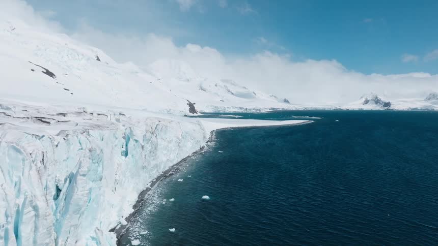 An aerial view drifts slowly along the towering textured ice wall of a massive glacier as it meets the deep blue arctic ocean with snowcovered mountains visible in the distance