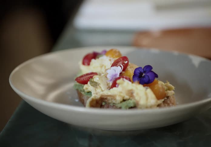 A dessert type of dish with cream and flowers decorating the top is on a table in a white bowl while a blurred person is in the background