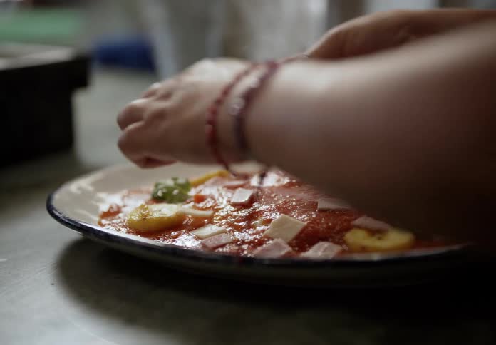 A persons hands delicately sprinkle fresh green herbs over a dish on a ceramic plate the dish appears to be a tomatobased savory preparation garnished with sliced bananas and pieces of white cheese
