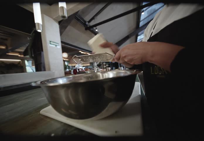 The video shows a closeup of a chef in a professional kitchen grating a block of white food likely cheese or another solid ingredient into a large stainless steel bowlthe chef is wearing a black apron and is standing at a counter with a white cutti