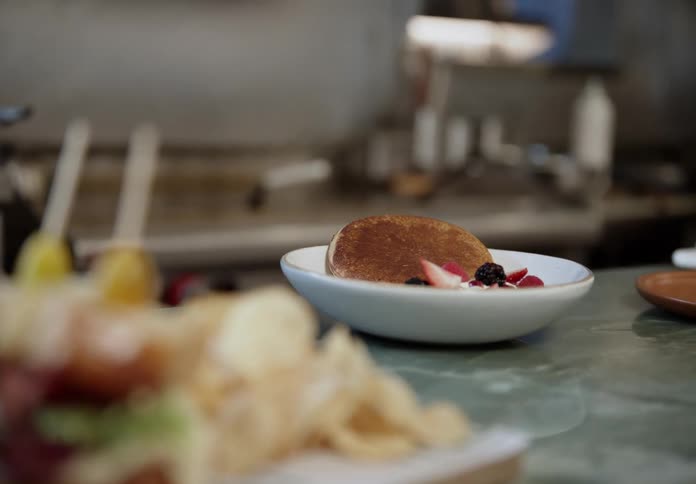A fluffy pancake topped with fresh berries rests in a white bowl on a countertop with another meal blurred in the foreground