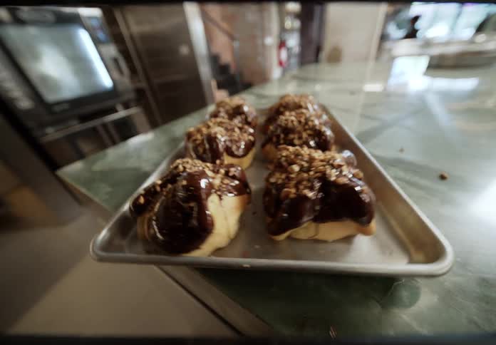 A hand places a small metal tray holding five large pastries onto a green marble counter the fresh buns are generously covered in a thick chocolate glaze and sprinkled with chopped nuts