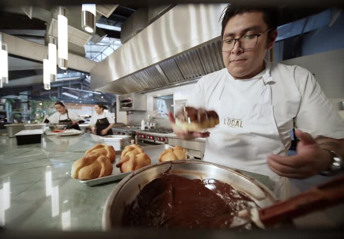 In a bustling professional kitchen a chef dips a pastry into a bowl of rich melted chocolate while other cooks work in the background