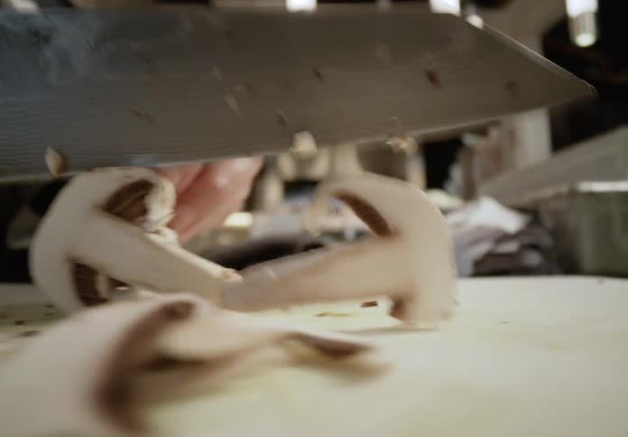 A chefs hand rapidly slices mushrooms on a cutting board in a busy modern kitchen