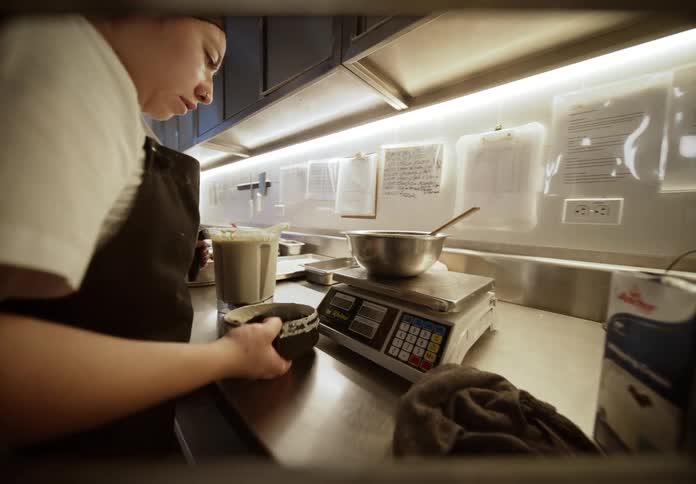In a busy commercial kitchen a female chef carries a blender pitcher to a prep station she then carefully pours the blended contents into a bowl set on a digital scale to weigh it