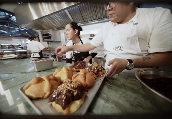 In a bustling professional kitchen a chef dips a pan de muerto into a bowl of chocolate before placing it on a tray and sprinkling toppings