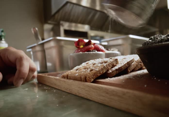 A persons hand sprinkles powdered sugar onto cookies on a counter in a restaurant kitchen setting