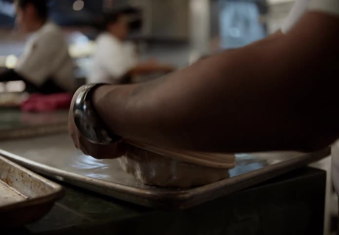 In a softly lit kitchen a baker carefully turns a round loaf of dough out of a proofing basket and onto a floured baking sheet