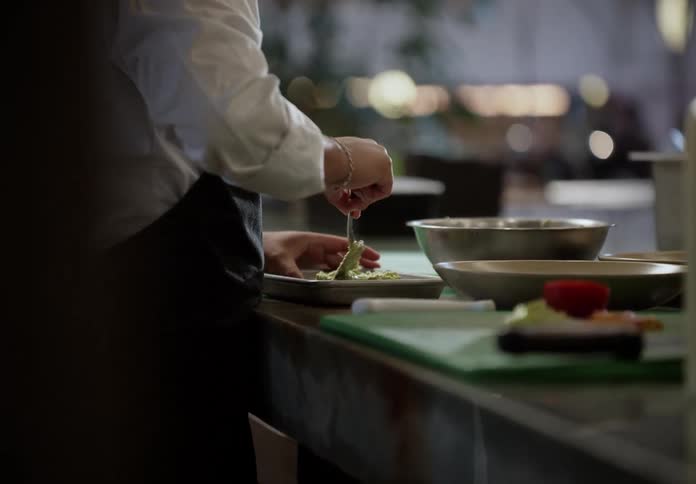 A chef wearing a white shirt and black apron is using a fork to carefully plate a portion of green food from a silver bowl the chef appears to be meticulously arranging the food on the rectangular serving dish at a kitchen prep counter