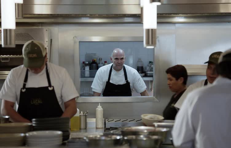 A professional kitchen where multiple cooks wearing white shirts and black aprons are busy working with stacks of bowls and ingredients in the foreground while the bald head chef is centrally framed in a stainless steel service window