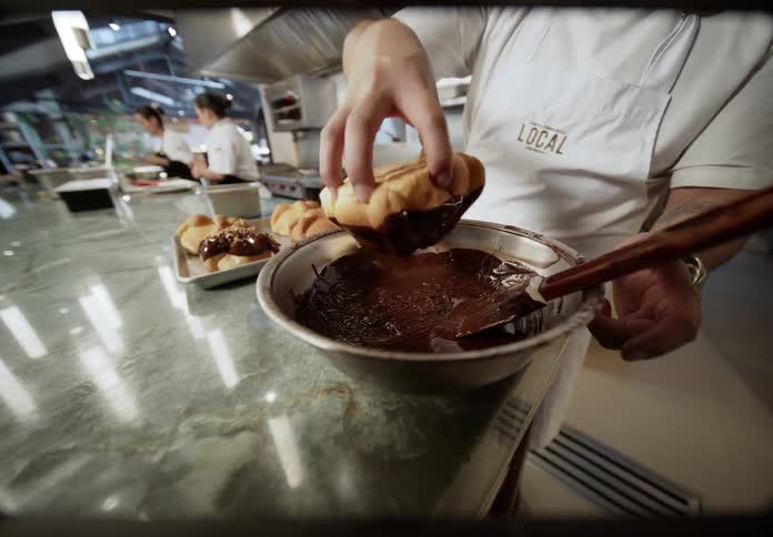 In a busy restaurant kitchen a chef dips pastries into chocolate and places them on a tray while others prepare food in the background