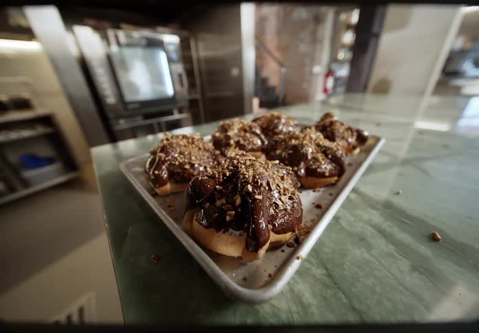 The video displays a tray of six freshly baked donuts or sweet buns generously topped with a rich dark chocolate glaze that drips down the sidesthey are sprinkled with crunchy nut pieces and sit on a metal baking tray which rests on a marbled green