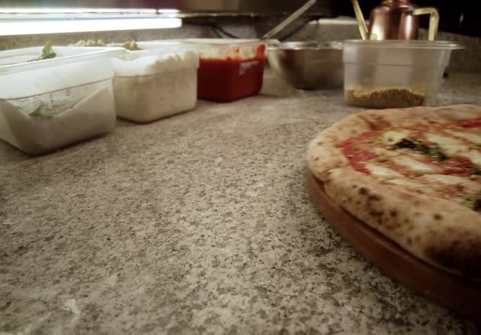 A freshly baked woodfired pizza topped with cheese tomato sauce basil and crushed pistachios rests on a wooden serving board a hand reaches in to slide the pizza board off the speckled granite counter where various preparation ingredients are store