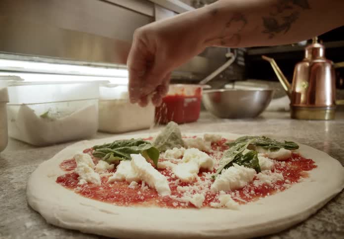 The video features a closeup shot of a raw pizza being prepared on a stone or flourdusted surface which already has a tomato base chunks of mozzarella cheese and fresh basil leavesa cooks hand visible in the upper frame is sprinkling fine