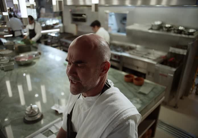 A chef standing in a professional kitchen wearing a white uniform and a dark apron as he briefly turns his head and talks to other workers and the cooking area where other kitchen staff in white uniforms are working