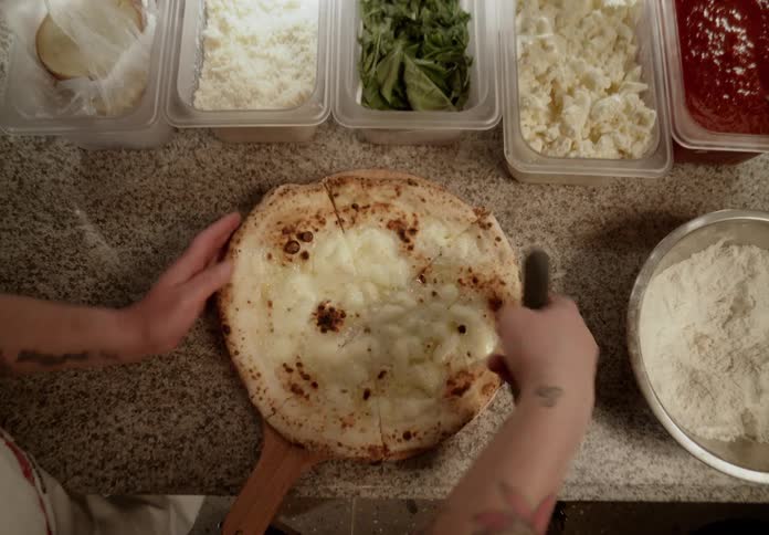 A tattooed cook slices a freshly baked cheese pizza surrounded by neatly arranged ingredients on a countertop
