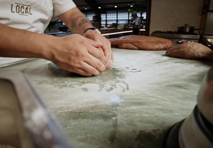 A cook in a restaurant kitchen with other employees around working is rolling dark dough on a counter top with flour
