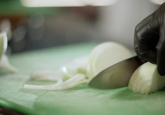A gloved hand uses a chefs knife to quickly slice a large half of a white onion the onion is being thinly cut on a green plastic cutting board in a closeup shot