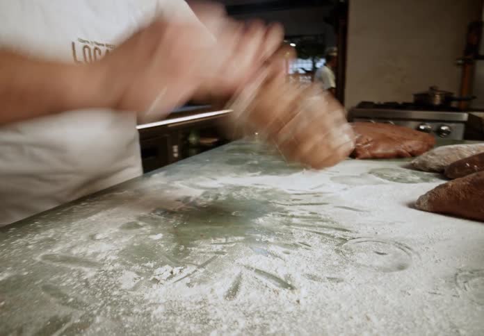 A middleaged man wearing glasses and a white apron uses his bare hands to prepare dough on a table inside a bakery at midday