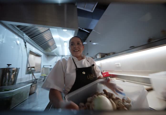 A smiling female chef walks through a busy commercial kitchen and stops at a prep station she picks up a large plastic container filled with mushrooms and carries it to another counter