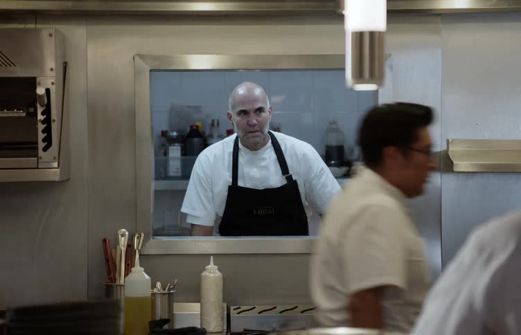 Workers and servers move through a restaurant kitchen as a chef watches through a window