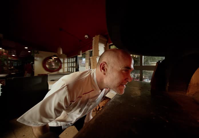 Male chef bends to stare into a lit fire burning oven in a commercial restaurant during the daytime