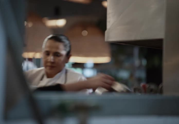 A pair of cooks move behind a kitchen pass partially obscured by equipment and warm hanging lights in the background