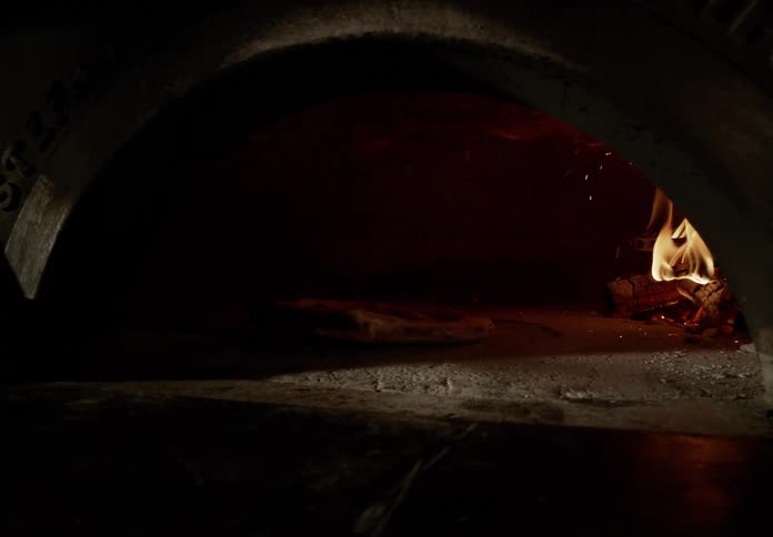 Inside a dark arched pizza oven a single pizza bakes on the hot stone floor to the right a pile of wood burns brightly its flames flickering and casting a warm glow